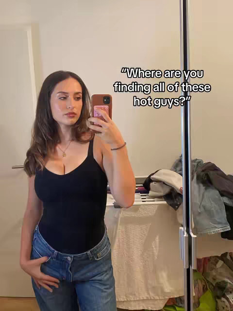A young woman takes a mirror selfie in what appears to be her bedroom. She is wearing a black tank top, blue jeans, and has long brown hair. Text overlay on the image reads, "Where are you finding all of these hot guys?"
