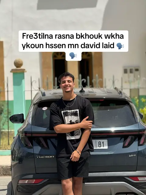 A young man stands in front of a dark blue SUV. He is wearing a black t-shirt and black shorts, smiling, and leaning slightly on the car. The background shows a building and the sky. Text overlay at the top reads in Arabic, with speech bubbles.