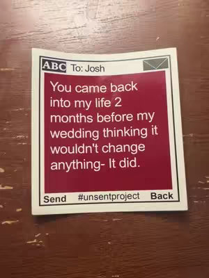 A square sticker on a wooden surface. The sticker has a white border with a red rectangle in the center. The text on the sticker says 'ABC To: Josh' followed by a message in white font. The background is a dark wood.