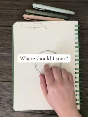 An overhead shot shows a notebook opened to a blank, off-white page. A hand is positioned over a sketched circle. Above the notebook, three pens in shades of green, light blue, and pink are laid out.