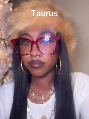 A woman with long black hair, wearing a fur hat and large red-rimmed glasses, a nose ring, and gold jewelry. The word "Taurus" is overlaid on the image.