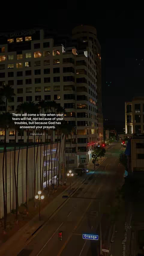 A nighttime cityscape featuring a high-rise building with lit windows and a street below. The sky is dark, and palm trees line the street, with the text of a religious quote overlaid.