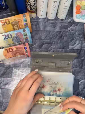 A top-down shot shows a person's hands opening a gray wallet filled with labeled envelopes. Euro banknotes (50, 20, and 10) lay to the left. The background has a patterned, dark gray brick or tile appearance. Items like some skincare products and a calculator lay on the right.