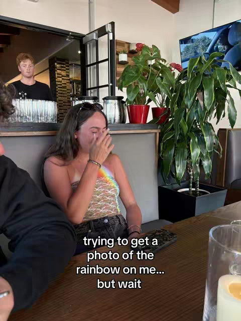 The cover image shows a woman sitting at a table indoors, seemingly at a restaurant or cafe. She has dark hair and is wearing a patterned strapless top. She is crying while her hand is covering her face, with a rainbow projected across her chest. Behind her, there's a man standing at a counter. The background includes interior elements like plants, a television screen, and kitchen equipment.