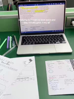 A top-down shot shows a laptop at the center, displaying highlighted text on its screen. A green desk is covered with pens, two notebooks, and a book labeled "USMLE Study Hacker". A printed sheet labeled 'Questions Solved' is visible. The scene is lit with natural light, suggesting a study environment.