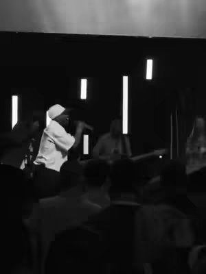 Black and white photo of a worship service. A person in a white shirt and hat sings into a microphone, a band is visible in the background, and the foreground features the silhouettes of seated audience members.