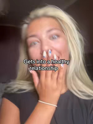 A young woman with long blonde hair is shown in a close-up, covering her mouth with her left hand. Her nails are manicured with white and blue polish. The background is blurred, with warm lighting. A text overlay that reads, 'Gets into a healthy relationship'. She wears a black top and a white bracelet on her wrist.