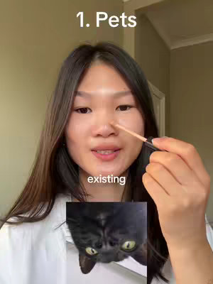 A young woman with long dark hair applies makeup while looking at the camera. Above her head is the text "1. Pets". Below her is an image of a black cat with wide green eyes. The background is a plain wall with a door frame visible on the right.
