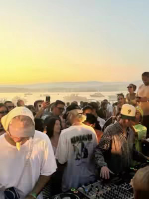 The cover image shows a crowd of people at an outdoor event, likely a music festival or party. In the foreground, there is a person wearing a white t-shirt and a yellow bandana. The background shows a crowd of people, with a view of the water and buildings. The image is taken at sunset as the sky is yellow and orange, the composition suggests motion and liveliness.