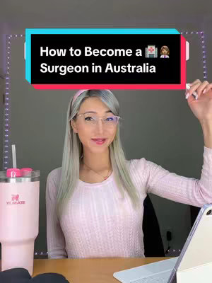 A selfie-style shot of a woman in a pink sweater, seated at a desk with a pink tumbler and a tablet. Behind her is a light fixture with small lights, creating a soft glow. The woman is smiling and appears to be looking at the camera. Overlayed on top is a black rectangular banner with white text that reads "How to Become a Surgeon in Australia".