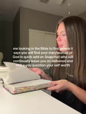 A young woman with long brown hair seated appears to be looking at a Bible in her hands, with pages open wide, while seated at a desk. The lighting is soft, and the room appears to be indoors.