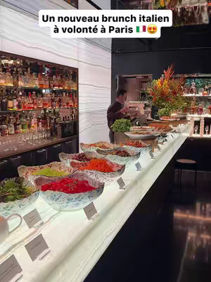 An interior shot of a brunch buffet at the Bvlgari Hotel Paris. Bowls of food are arranged on a white lit counter. A bar with numerous bottles can be seen in the background. A person is preparing or arranging food at the end of the buffet.