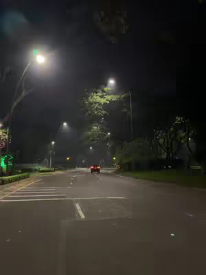 A nighttime shot of a road receding into the distance with a car's taillights visible. Streetlights illuminate the scene, and trees line the sides of the road under a dark sky.