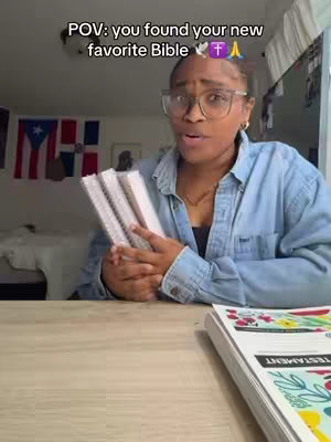 A woman with glasses is holding multiple books and looking at the camera. The background includes a room with flags.