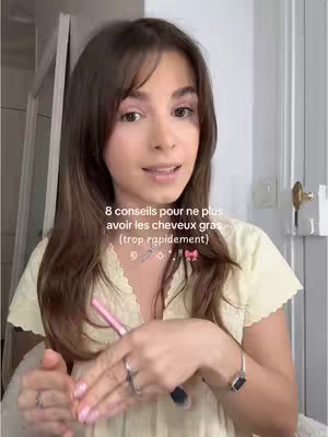 A young woman with long brown hair and bangs is centered in the frame, facing the camera. She is wearing a light yellow top with a detailed neckline. The background appears to be a neutral-colored wall with some visible fixture. The woman is holding a pink makeup applicator to her lips. Above her, a text overlay states the video's topic, and icons are interspersed around the text.