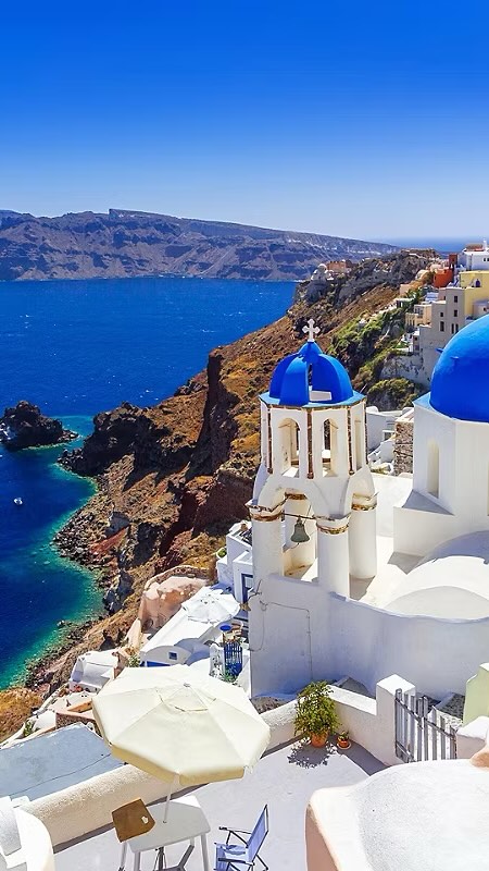A vibrant image showcasing a picturesque coastal scene in Santorini, Greece. The primary focus is on the iconic blue-domed churches set against white-washed buildings, perched on a cliff overlooking the Aegean Sea. The foreground features a partially visible patio with a table, chairs, and a sun umbrella, suggesting a viewpoint from a cafe or balcony. The composition is bright and sunny, with a clear blue sky and deep blue waters creating a strong contrast with the crisp white and blue architectural elements. The overall impression is one of idyllic beauty and tranquility.