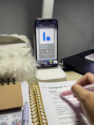 A person is studying at a desk, with an open notebook, pens, and a phone playing a podcast or audio. The scene is lit with a desk lamp and has a cozy, organized look.