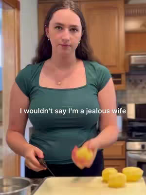 A woman stands in a kitchen setting. She wears a green top and dark pants, holding a knife and a partially peeled apple. The background features kitchen cabinets and appliances, giving the image a domestic focus.