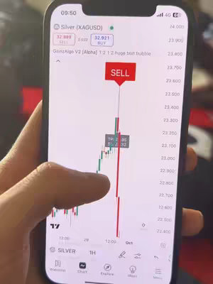 A person is holding a smartphone displaying a trading application, with a chart and price action data. The phone's screen shows a green candlestick chart trending upwards, with a red 'SELL' indicator overlaid on the chart.  The phone is held against a dark-colored background. A finger is touching the screen indicating some kind of real-time interaction with the app.