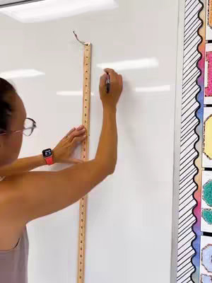 A woman is shown from mid-torso up, standing in front of a white dry-erase board. She is wearing glasses, a light colored tank top, and a red smart watch. She holds a ruler and pen in front of the board, as if measuring and marking it with the pen.
