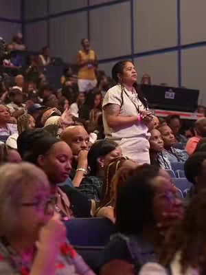 A crowd of people in an auditorium setting. The image is composed of people, with the central focus on a woman standing up in the crowd.  She has a white top and is looking up. There are other people in the audience, who appear to be listening attentively. The background is a light-colored wall. The overall tone of the image is one of a religious meeting.