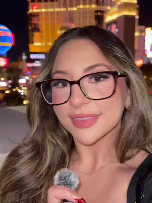 A woman with long, wavy hair and glasses is centered in a selfie with a city backdrop at night. She holds a furry microphone in her hands and smiles slightly. The background features bright city lights and tall buildings.