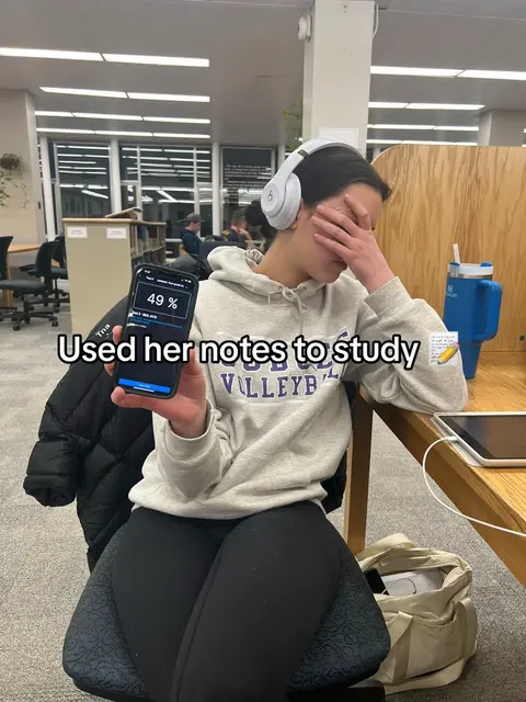 A person is holding a phone displaying a studying app, while sitting at a table in a library setting. The person has their hand on their face and is wearing headphones.