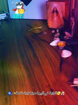 The image shows a close-up, low-angle shot looking down at a wooden surface decorated with items related to tarot readings. There's a golden compass, a small lit candle, a seashell, and small figurines scattered on the surface. In the upper left corner is a cartoon bear character with an Aries symbol in a shield. Behind the bear, a black and white design creates a backdrop, and there's a book or container towards the right with a picture on it. The image uses a purple hue with highlights, creating a mystical feel, suggesting a tarot or astrological theme.
