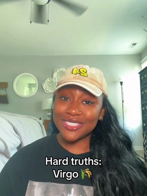 A woman wearing a baseball cap smiles at the camera in a well-lit room, with text overlayed at the bottom reading 'Hard truths: Virgo'. The background includes a bed, a decorative mirror, and a ceiling fan.