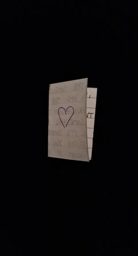 An image of a small, folded card with a drawn heart on it, set against a dark background. The card appears to be made from white or off-white paper, with visible lines indicating its folds. The heart is drawn in a dark color, likely purple or blue, and is centered on the card. Handwritten text is visible on parts of the exposed paper. The background is completely dark, which emphasizes the card. The overall composition is simple and intimate.