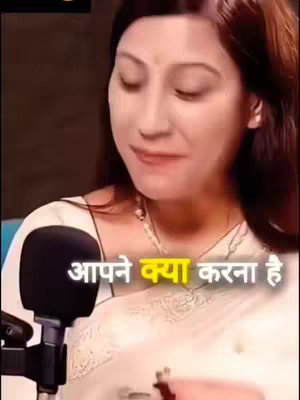 A woman is seated facing the camera, with a microphone positioned in front of her. She is wearing a white outfit and a necklace with a pendant around her neck. Text in Hindi is overlaid at the bottom, with a yellow highlight on some of the words.