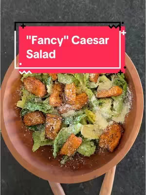 The image shows a top-down view of a Caesar salad in a wooden bowl. The red rectangle with white text overlay near the top of the image says '"Fancy" Caesar Salad'. The salad itself features a bed of green lettuce topped with golden-brown croutons.  Sprinkled across the top are flakes of what appears to be grated cheese. The food is set against a dark background.