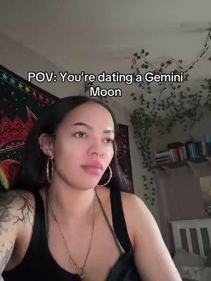 A woman with long dark hair and hoop earrings is centered in the frame, looking thoughtfully to the left. She wears a black tank top and layered necklaces. The backdrop is a room with a tapestry, decorative vines, and a glimpse of a bookshelf. Above the woman, text overlays read, 'POV: You're dating a Gemini Moon'.