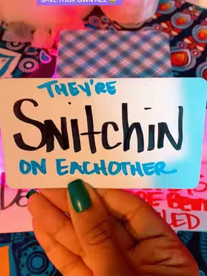The image shows a hand holding a white card with the words "THEY'RE SNITCHIN ON EACHOTHER" written in black and blue marker. The hand has long nails painted in a teal green color. The background includes tarot cards, crystals, and some blurred items and a teal background.