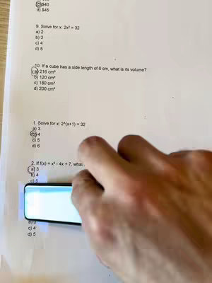 The image shows a close-up of a hand holding a digital pen over a piece of paper with math problems. The hand is centered, and the pen is positioned directly above the paper, ready to interact with it. The paper itself is white with printed questions and multiple-choice answers.