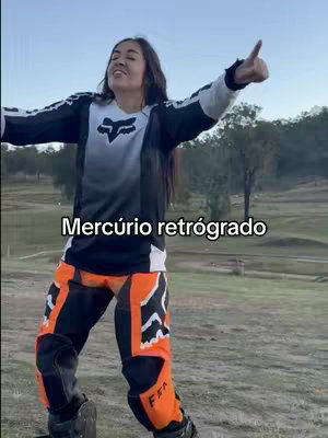 A woman wearing a Fox racing jersey and pants is standing on a grassy area with her arms outstretched and a thumbs up. The background features a green landscape with a clear sky. Text overlay reads 'Mercúrio retrógrado'.