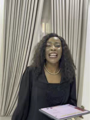 A woman with long, curly dark hair and a gold necklace is standing in front of a beige curtain. She is wearing a black dress and holding a tablet. She is speaking passionately, her mouth open and her facial expression conveying excitement or fervor.