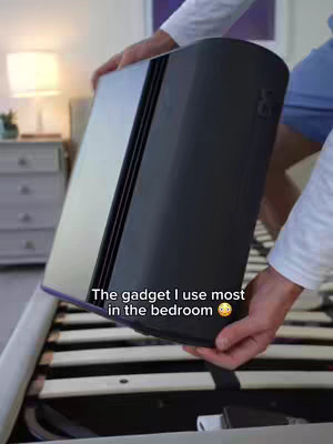 A person is holding a black rectangular device, possibly a smart device or a tech gadget, positioned over a bed frame with a slatted base. The background is a bedroom setting with a lamp, a nightstand, and a glimpse of a window or artwork. The scene suggests a focus on technology within a home environment.
