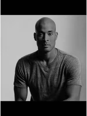 A black and white image of David Goggins, a bald man with a serious expression, wearing a gray t-shirt. He is centered in the frame, with a plain white background and a solid black bar at the bottom, presumably for aesthetic or branding purposes.
