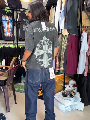 A man stands in what looks like a clothing store, viewed from the back. He's wearing a dark gray t-shirt with a white graphic and jeans, and he's looking to the left. Clothing racks and items are visible in the background, suggesting a store environment.