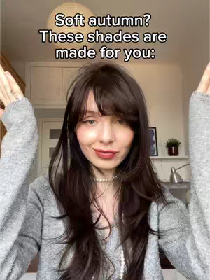 A woman with long brown hair and a soft, grey sweater stands with arms open wide, framing her face. Text overlays read, 'Soft autumn? These shades are made for you:' The background suggests an interior with neutral tones, consistent with the color palette.