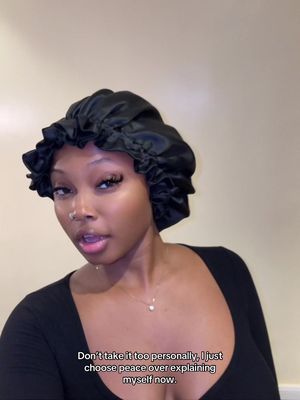 A woman with dark skin and long lashes looks toward the left. She is wearing a dark satin bonnet on her head and a simple necklace. Text appears on the lower portion of the screen.