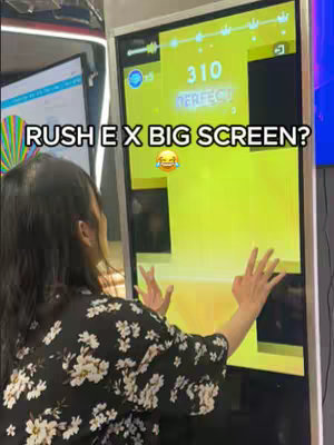 A woman is playing a rhythm game on a giant screen. The screen shows a yellow game interface with notes and a score of 310 'PERFECT'. The woman is seen from behind, wearing a floral patterned shirt, and her hands are raised in front of the screen. Text overlays the screen.
