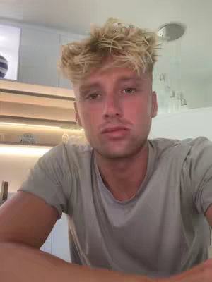 A young man with light-colored, styled hair is centered in a selfie-style shot. He's wearing a gray t-shirt, and the background shows a glimpse of a modern kitchen setting. His expression is neutral, and the lighting is soft.