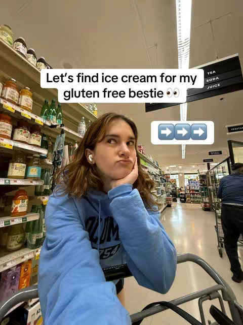 A woman with brown hair and in-ear headphones is in a grocery store aisle. She is holding her chin with her hand and looking up and to the side with a thoughtful expression. The aisles are filled with shelves of canned goods and other groceries. Text overlays at the top say, 'Let's find ice cream for my gluten free bestie' and directional arrows are in a box below.