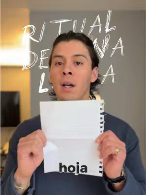 A man with dark hair holding a white sheet of paper opened with his hands, looking towards the camera. The background is a plain gray wall. Over the image, the handwritten text "Ritual de Luna Llena" is scribbled.