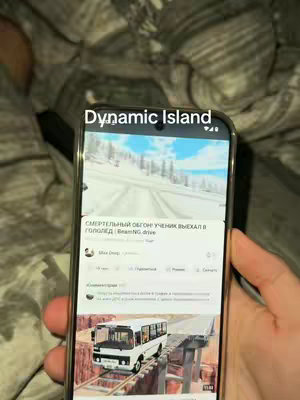A person is holding a smartphone displaying a video from YouTube. The phone shows a snow-covered road, a bus on a bridge, and the title "Dynamic Island" overlaid at the top. The background includes blurred bedding.