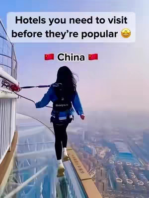 A woman is walking along a glass walkway high above a cityscape. The image is shot from behind the person, and the focus is on the daring nature of her placement in an expansive landscape. The sky is a gradient of blue and white, and the city below appears to be a modern metropolis.