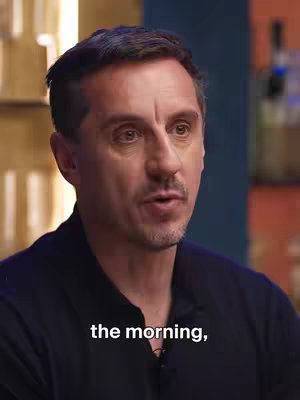 A medium shot of a man with short, dark hair and a somewhat stern expression. The background shows a warm-toned, somewhat cluttered setting. The man is wearing a dark blue or black collared shirt.