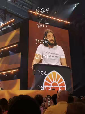 A man with a beard wearing a white t-shirt stands at a podium in front of a large screen. The screen has a light brown or orange background, with the words of the hook written on it. Many people are visible in the bottom of the view, presumably an audience.